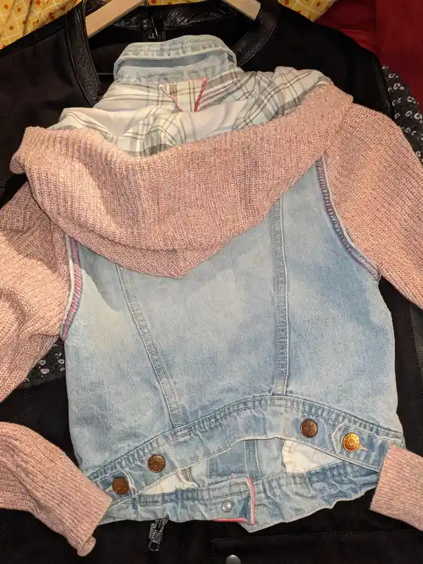A close-up photo shows a stack of clothes on a hanger, with a light blue denim vest on top, partially covered by a pink…