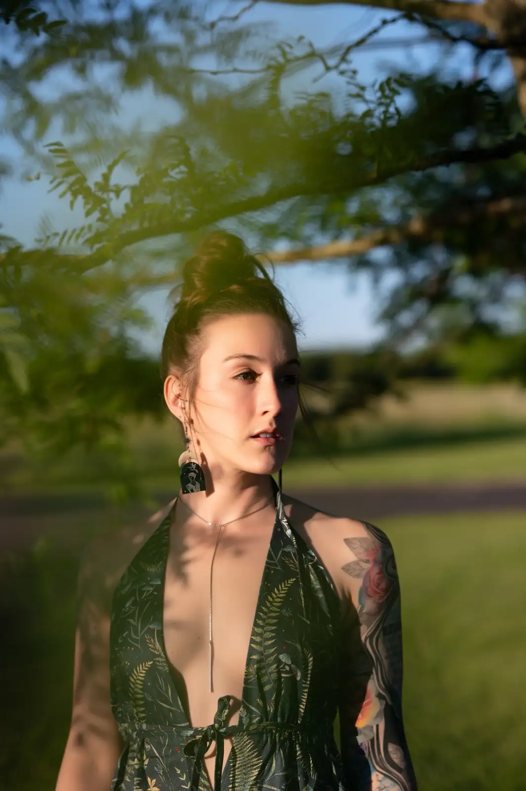A woman with a tattooed arm and leaf-patterned dress stands outdoors under tree branches, looking pensive. The overlaid text…