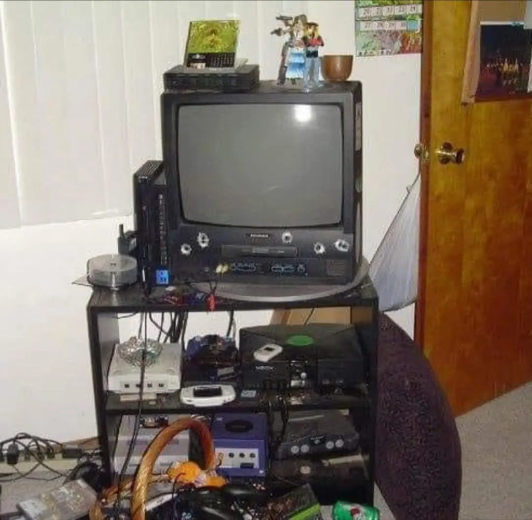 A cluttered, multi-tiered TV stand is packed with old gaming consoles, including a PlayStation, GameCube, and various other…