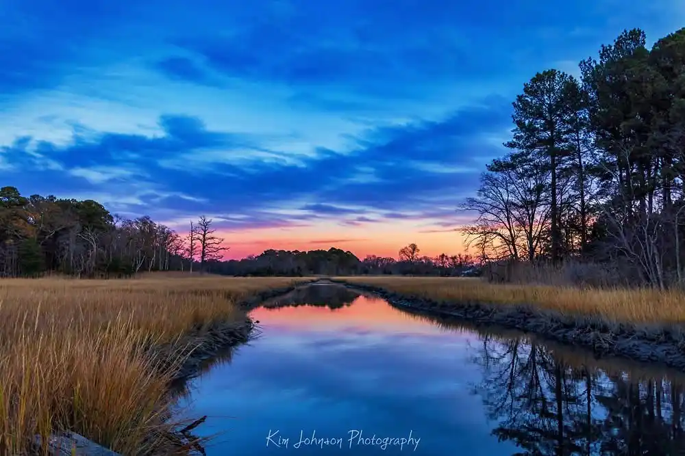 A serene landscape photograph of a narrow waterway flanked by tall golden grasses and trees, reflecting a vibrant sunset sky…