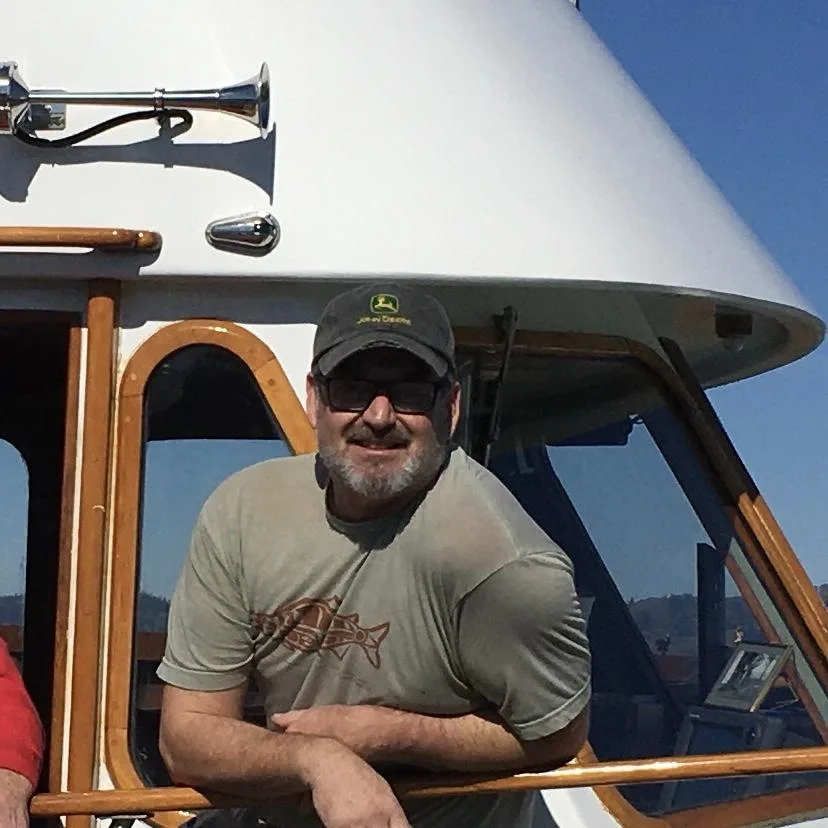 a man with a beard, sunglasses, and a John Deere cap leans on a wooden railing of a white boat with a horn on top. He smiles…