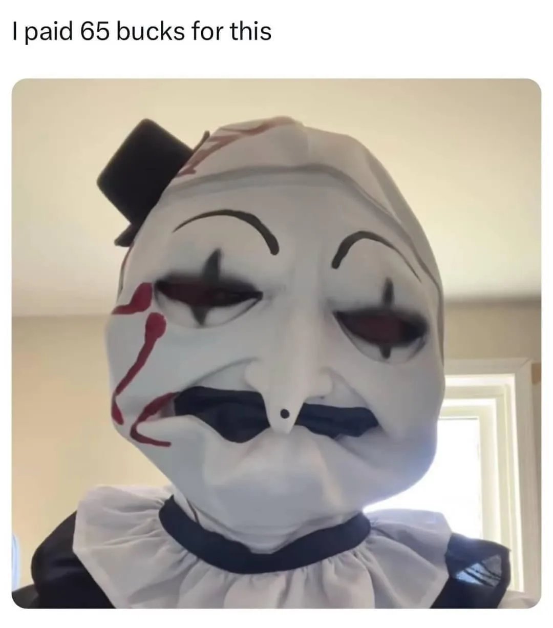 A person wearing a white, cartoonish clown mask with black eyes, a black mustache, and red paint resembling blood streaks on…