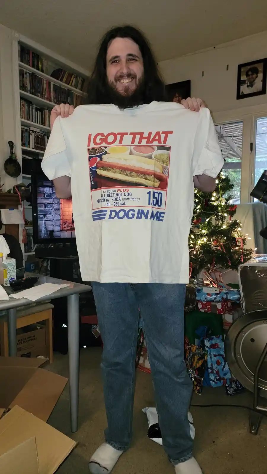 A man with long hair and a beard stands in a living room holding up a white t-shirt. The shirt features a vintage-style hot…