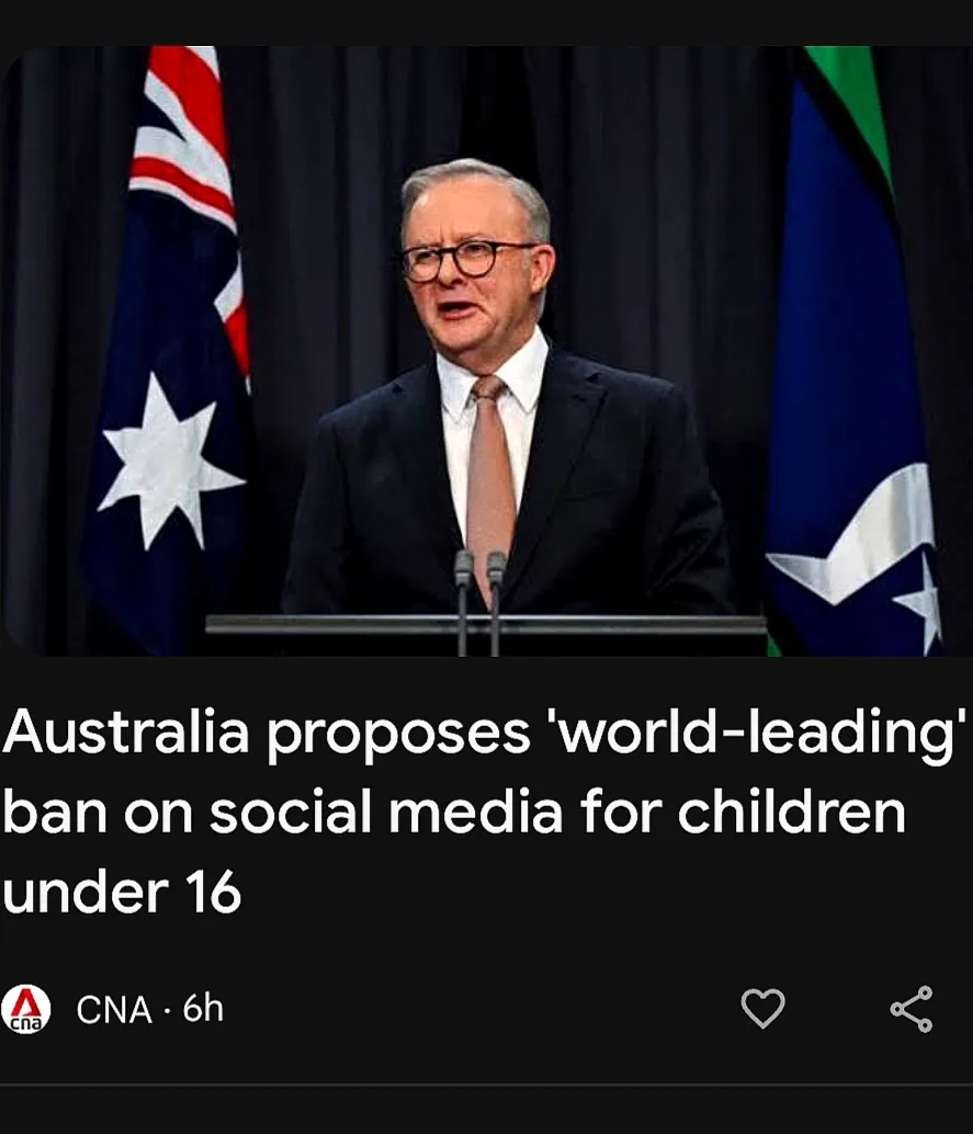 A man in a suit and glasses stands at a podium with two flags behind him, one being the Australian flag. The image is…