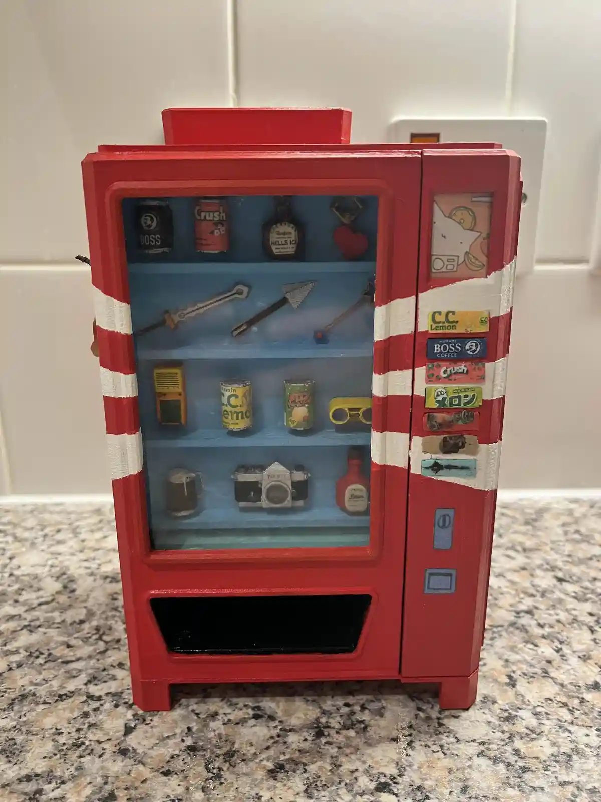 A small, red and white striped miniature vending machine sits on a granite countertop, its glass door open to reveal shelves…