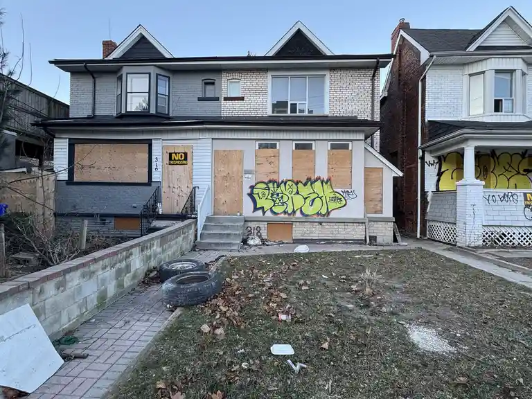 A dilapidated two-story house with all windows and doors boarded up with plywood, covered in bright green graffiti, and…