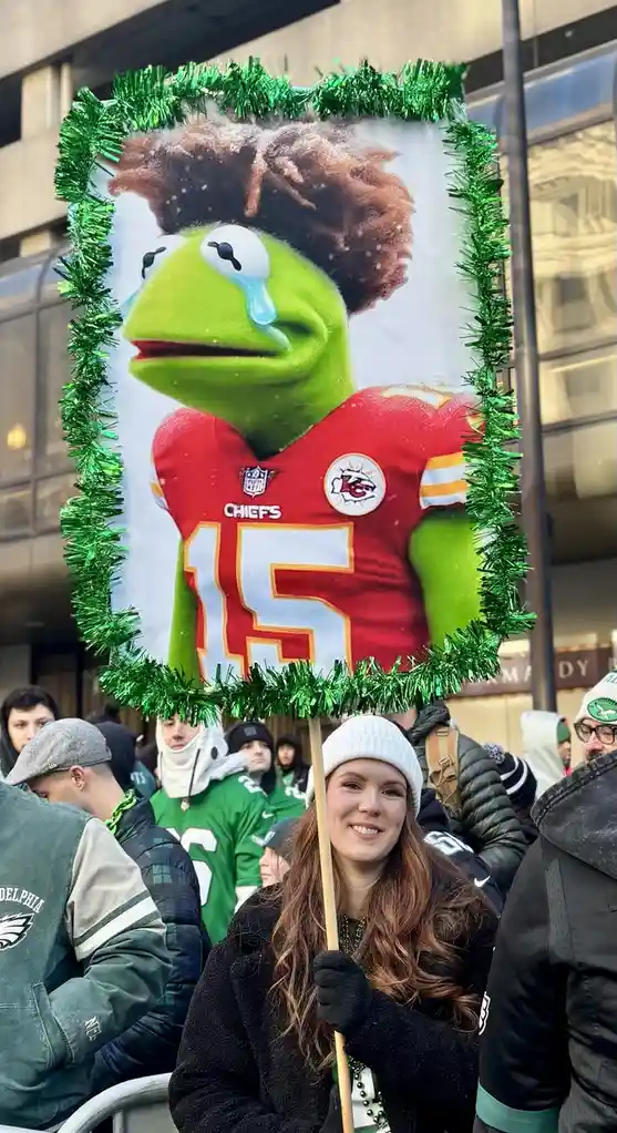 A person holds a decorated sign featuring Kermit the Frog with a crying expression, digitally edited to wear a red Kansas…