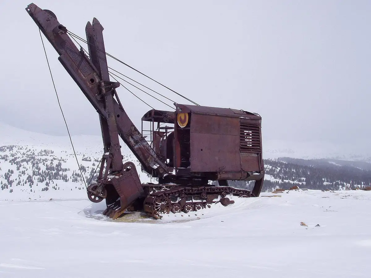 A rusted, abandoned tracked mining excavator sits partially buried in deep snow in a snowy mountain landscape. The machine…