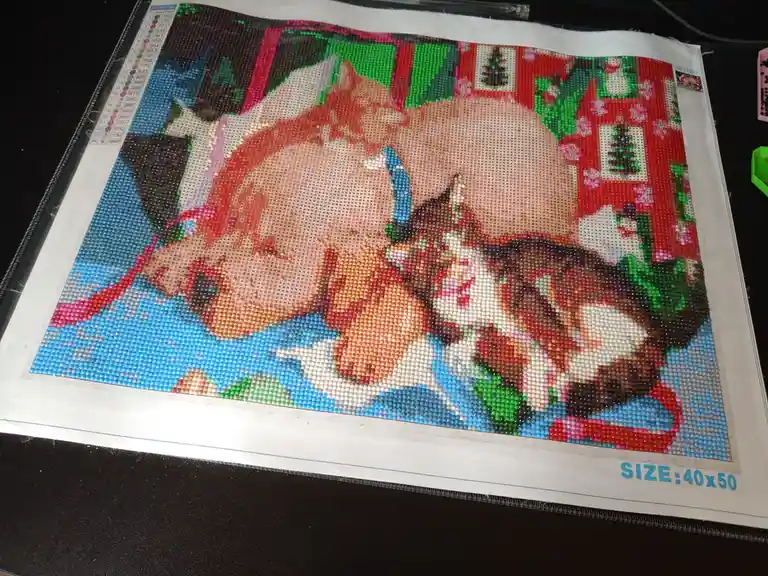 A partially completed diamond painting of a cat wearing a Santa hat, set against a festive red background with Christmas…