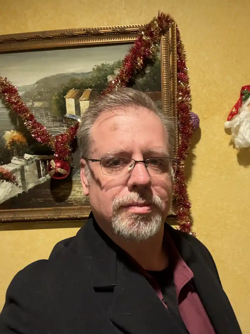 A middle-aged man with glasses and a gray beard stares directly at the camera with a neutral expression. Behind him, a large…