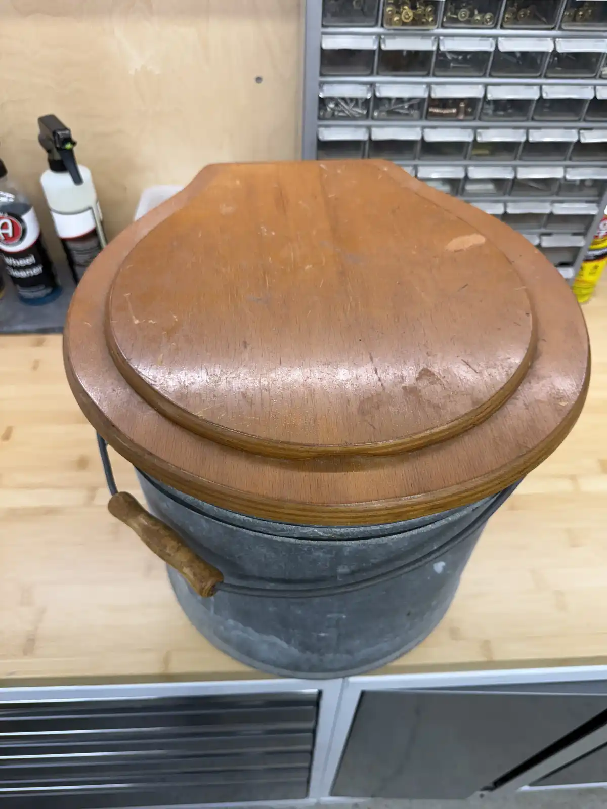 A wooden toilet seat lid is shown resting on top of a galvanized metal bucket with a wooden handle, placed on a workbench in…