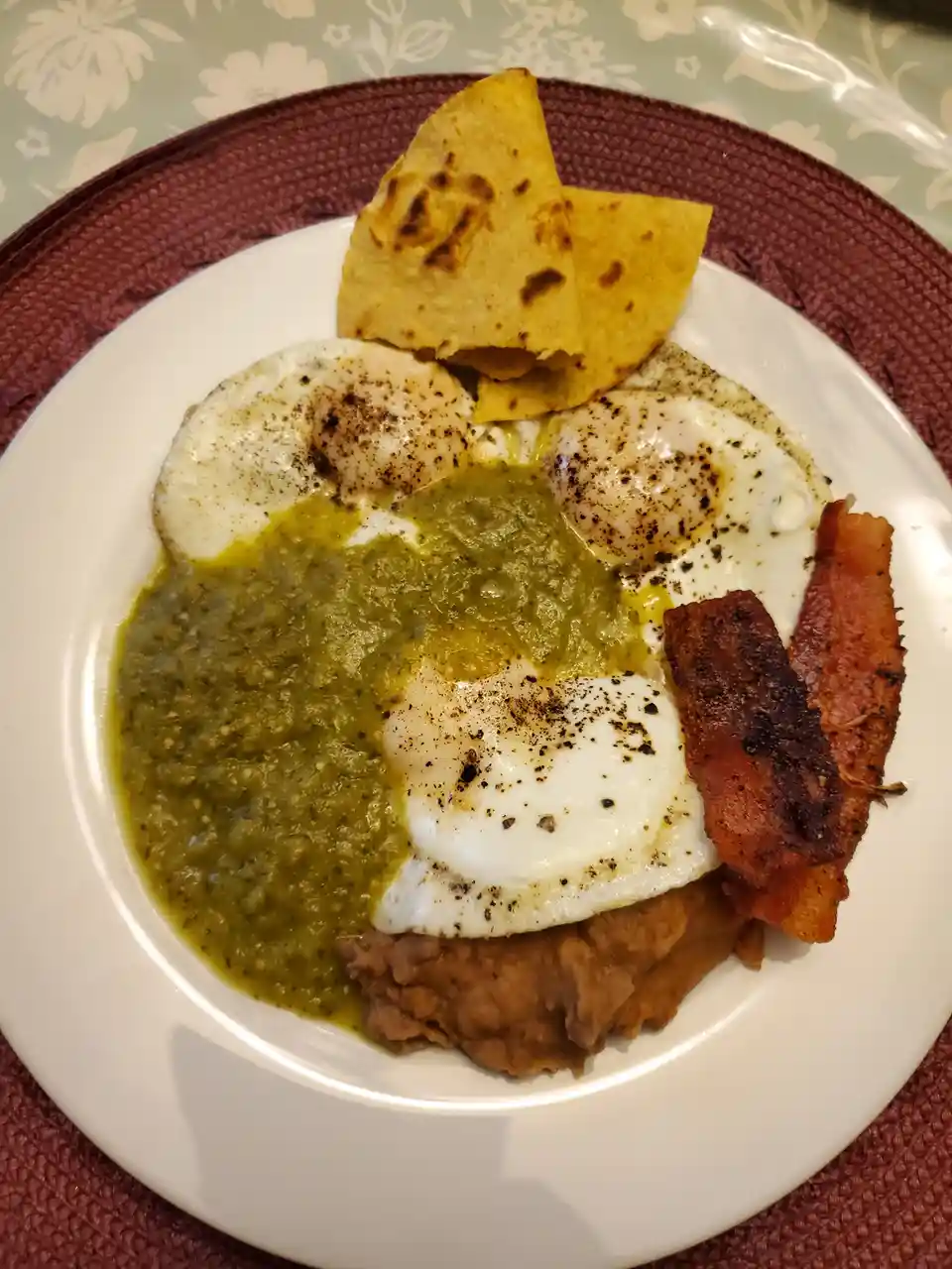 A plate of food featuring fried eggs, bacon, refried beans, and green salsa, topped with tortilla chips, with the text 'this…