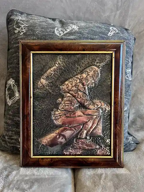A framed metal relief artwork depicts a seated figure with a large, textured, multi-limbed creature resembling an octopus or…