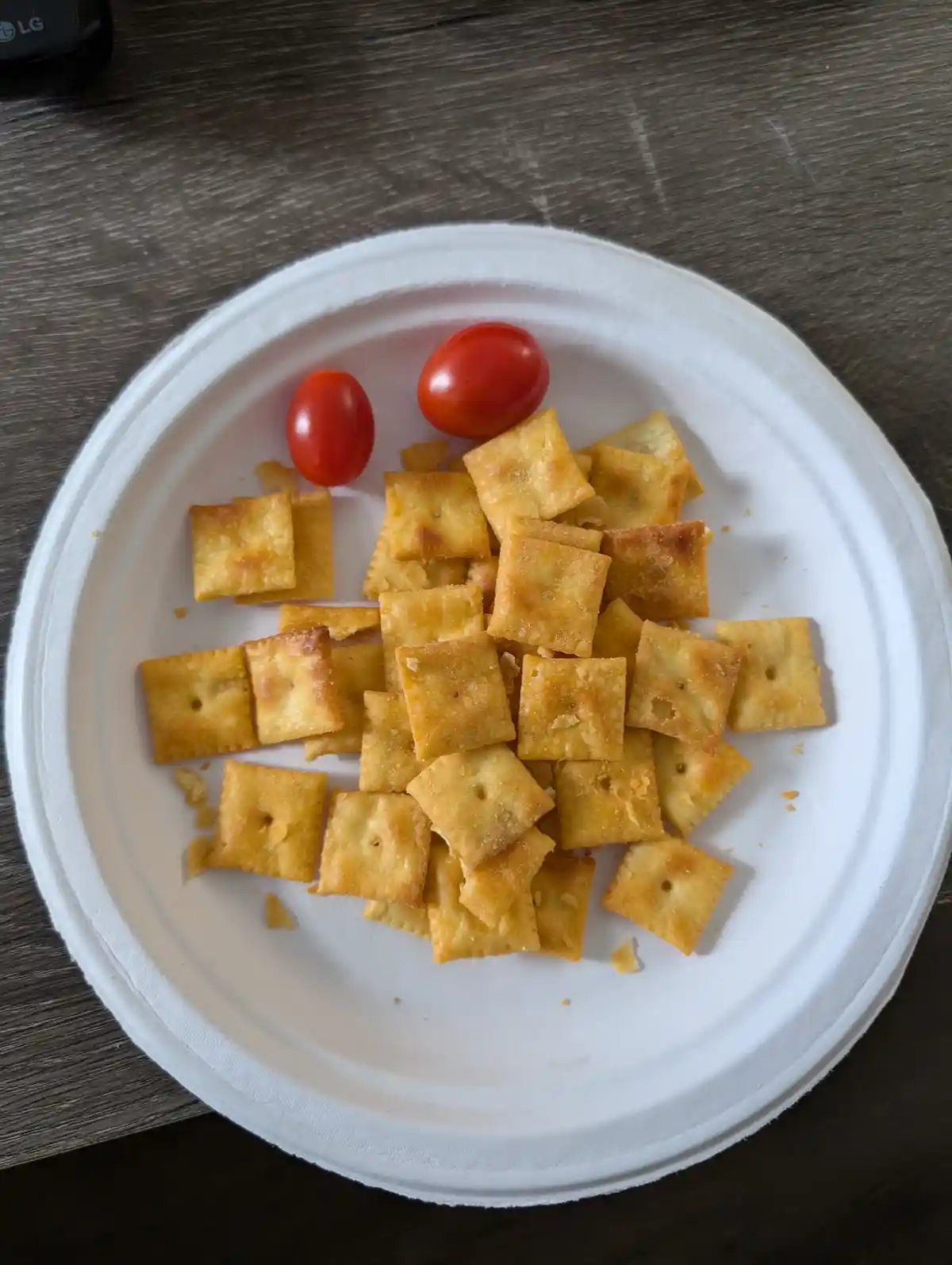 A white paper plate holds a pile of square, golden-brown crackers and two cherry tomatoes, arranged on a dark wooden table…