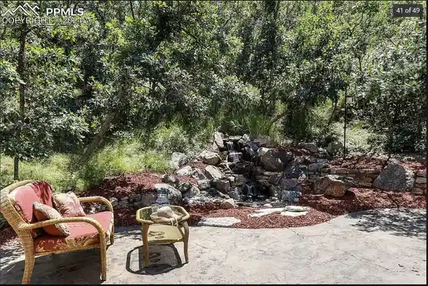 a real estate photo shows a backyard patio with wicker furniture and a small water feature made of stacked rocks and mulch…