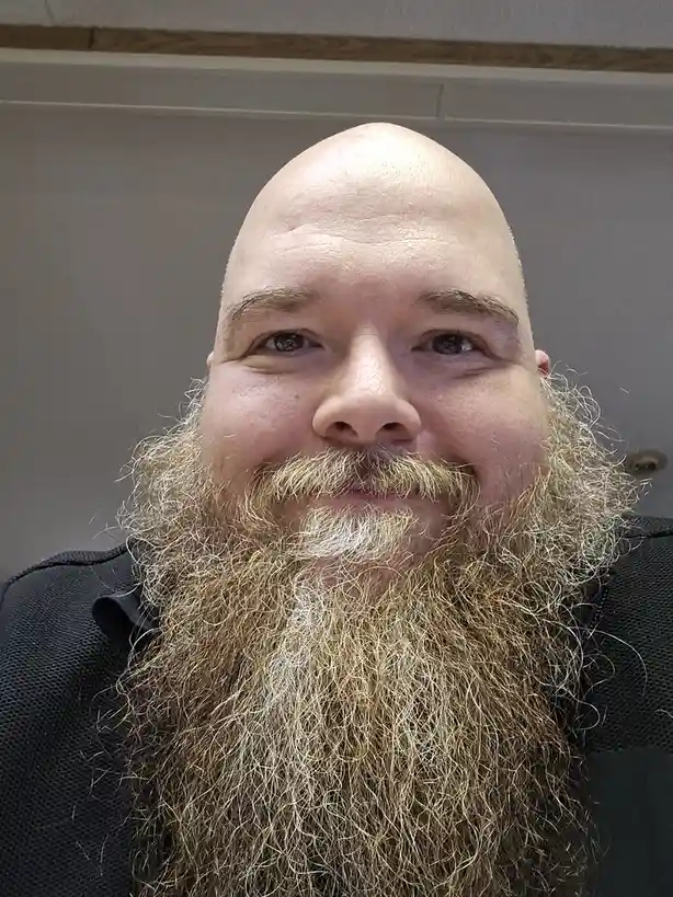 A close-up selfie of a bald man with a very long, full, and curly beard that is mostly brown with some gray hairs, covering…