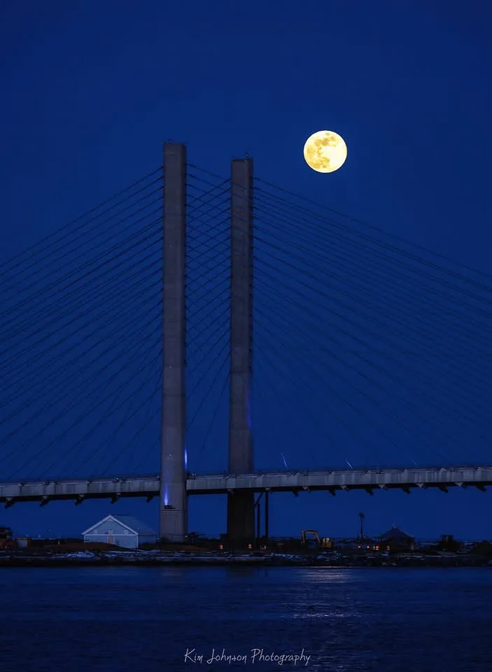 A full, bright yellow moon hangs in a deep blue night sky, positioned directly above the tall towers of a modern…