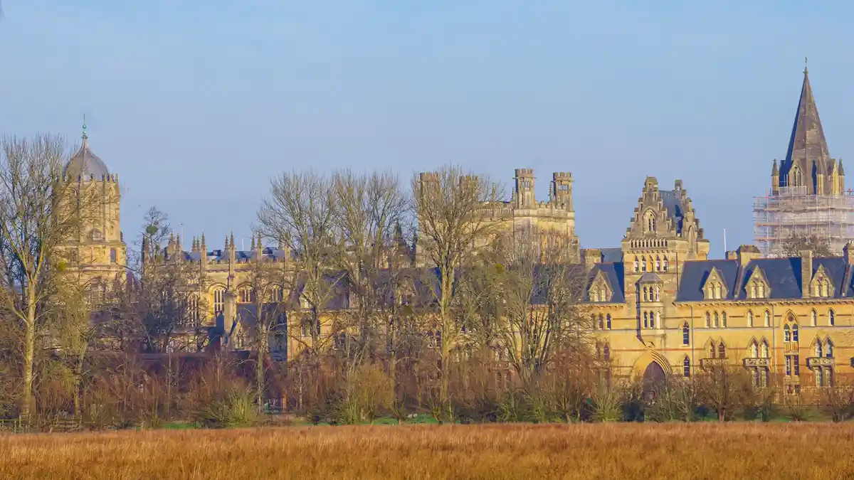 A wide shot of a grand, historic university campus with Gothic-style buildings, turrets, and spires, partially obscured by…