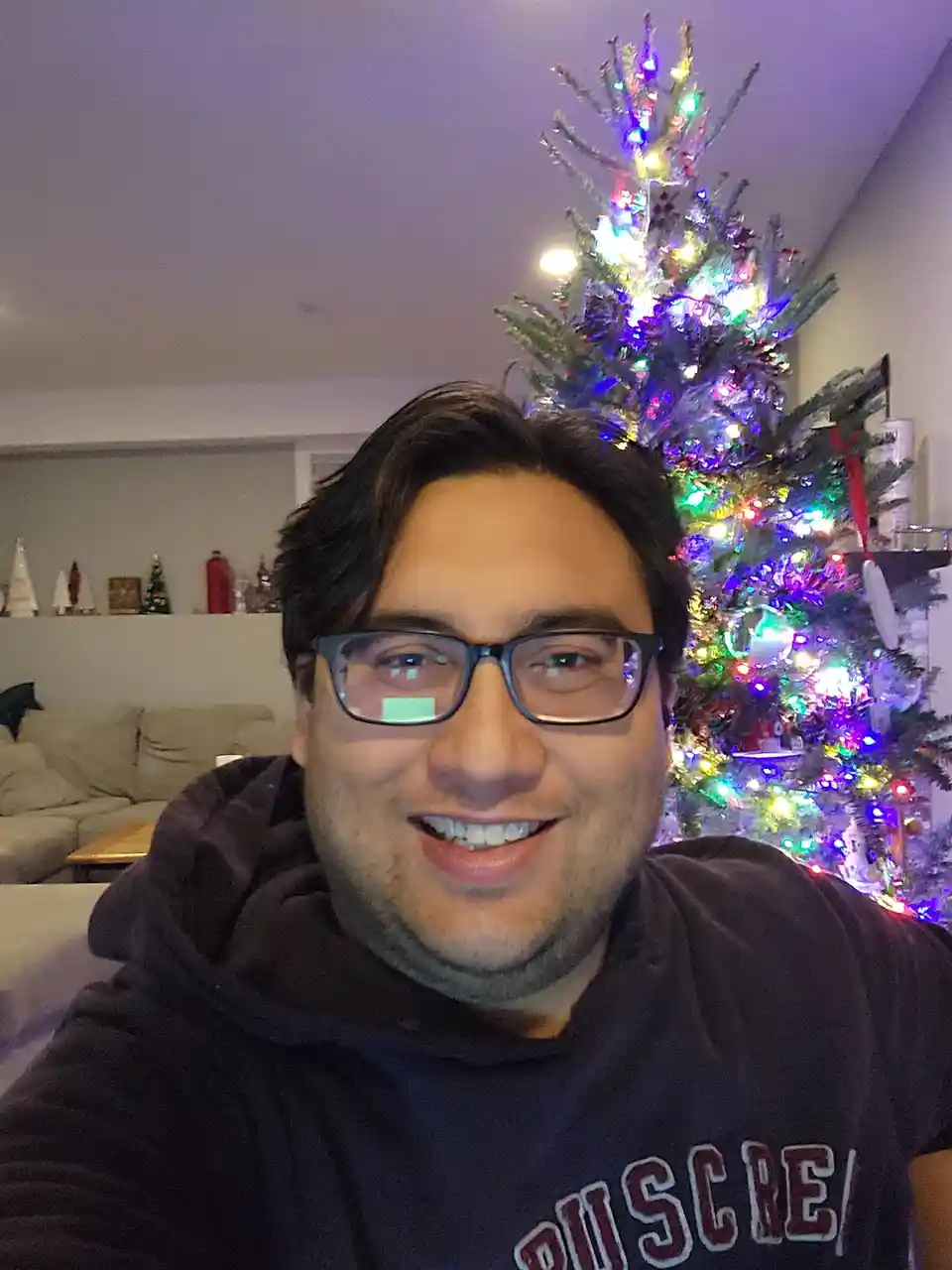 A man with glasses and a dark hoodie smiles at the camera while standing in front of a brightly lit, multi-colored Christmas…