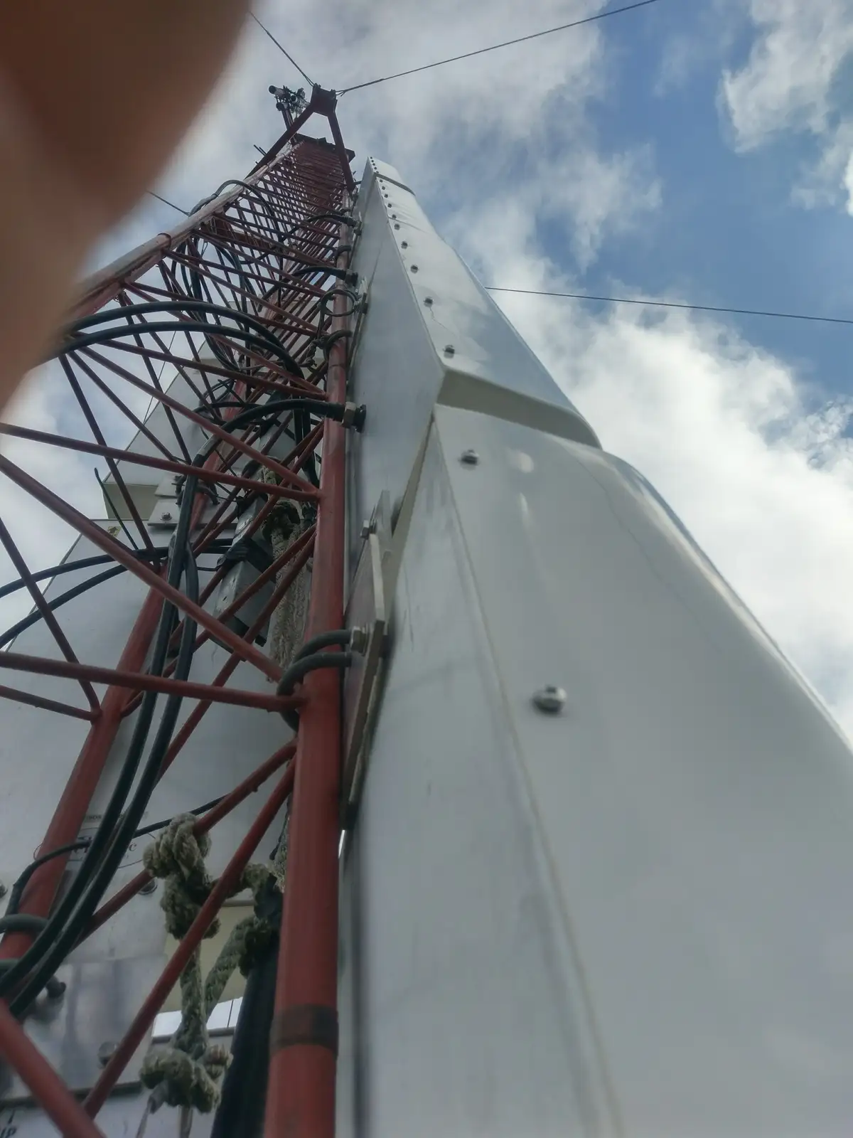 A low-angle photo looking up at a tall, white telecommunications tower with a red metal ladder and black cables running up…