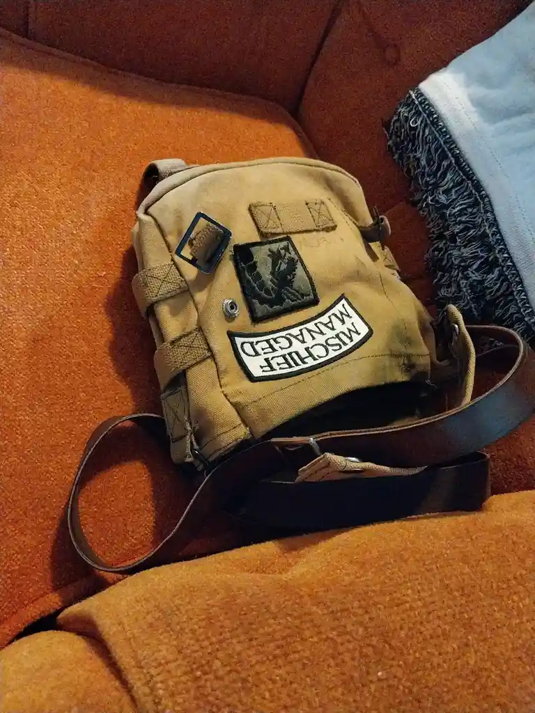 A tan tactical-style backpack with multiple patches rests on an orange couch. A prominent white rectangular patch on the…