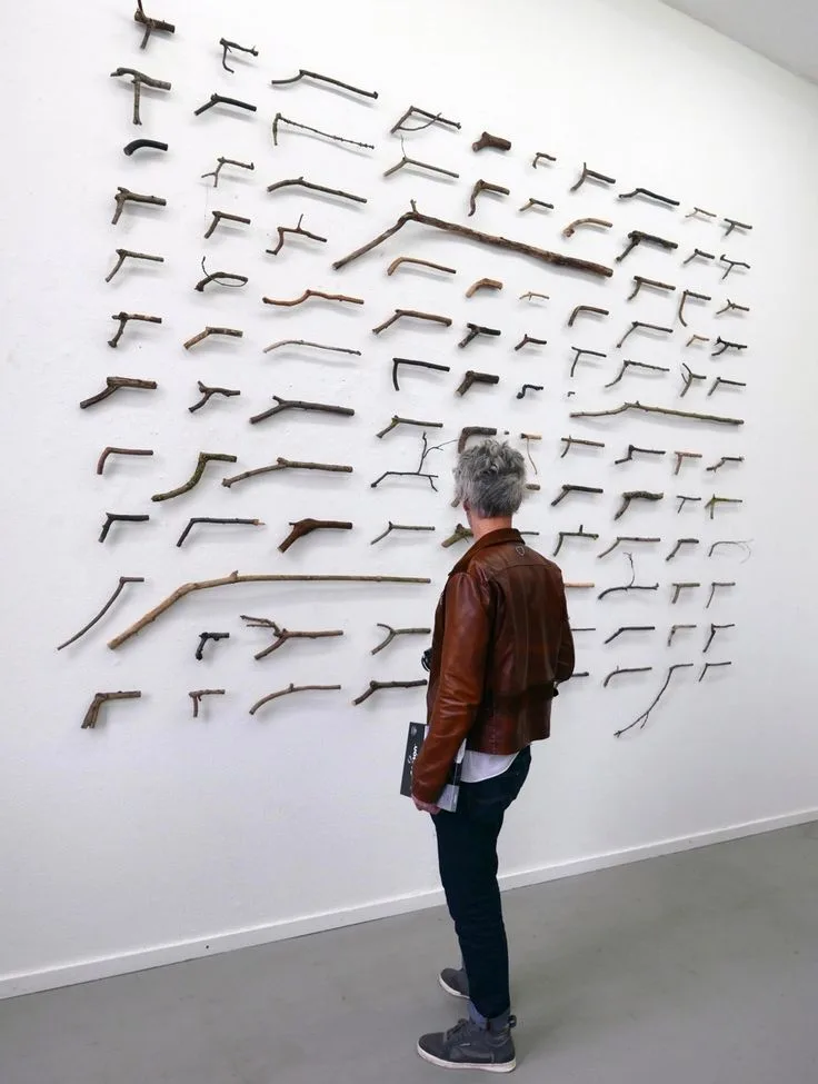 A person with gray hair wearing a brown leather jacket stands before a white gallery wall displaying numerous sticks…