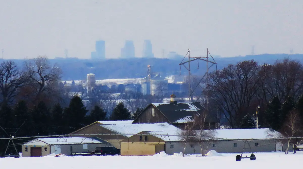A snowy rural landscape with farm buildings in the foreground, bare trees, and a distant city skyline visible through…