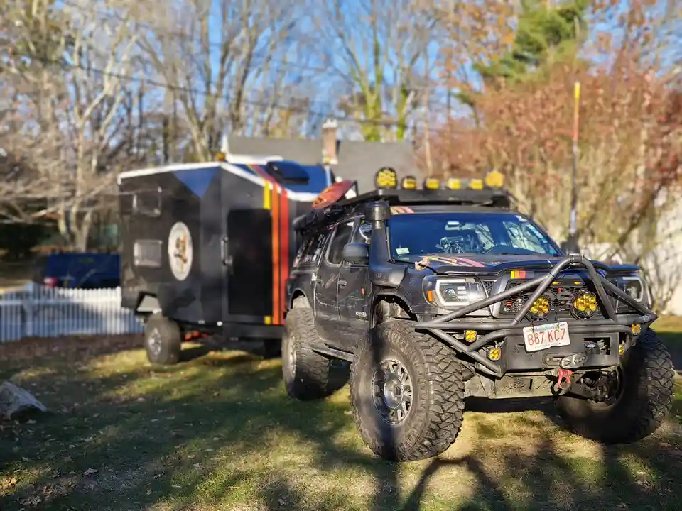 A heavily modified black off-road truck with large tires, a front bumper guard, and roof-mounted lights is parked on grass…