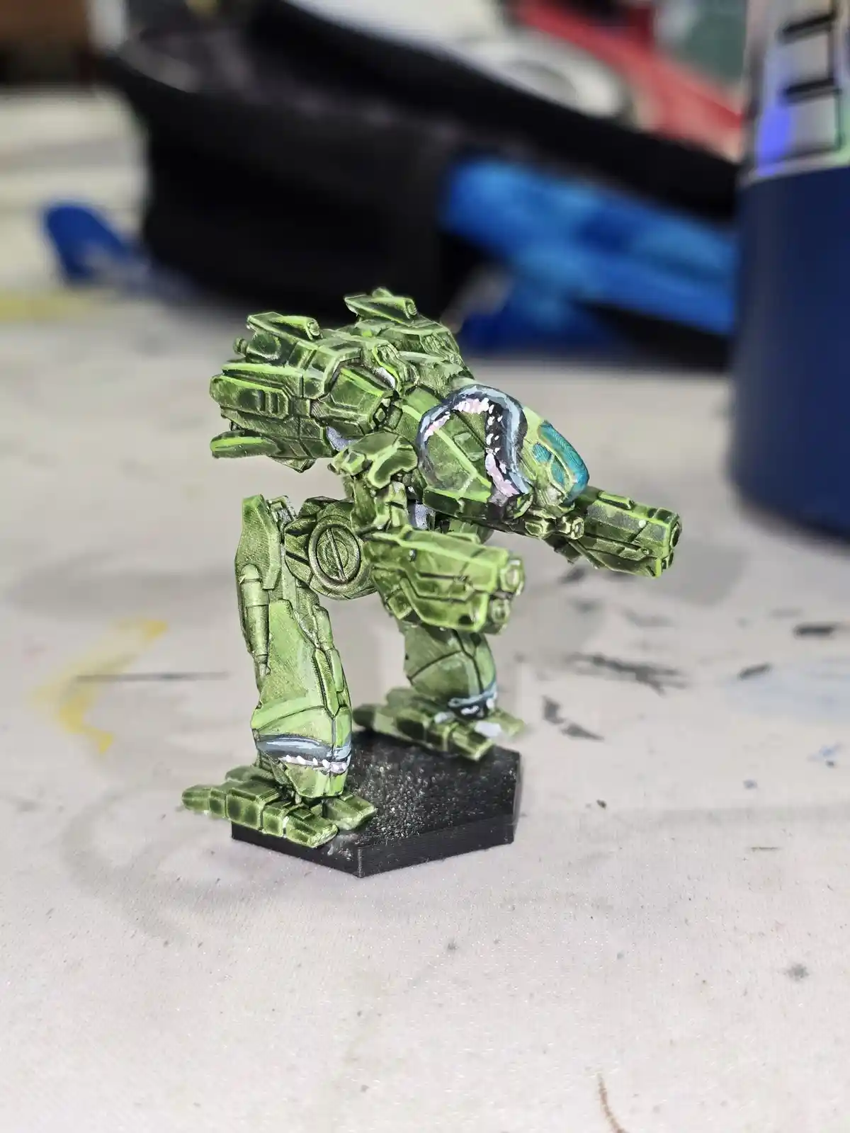 A green-painted miniature mech model stands on a black hexagonal base, its head and chest area painted to resemble a shark…