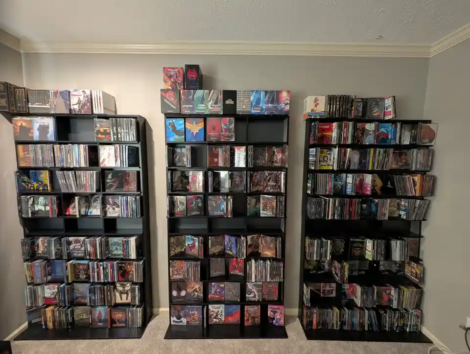 Three tall black shelves in a room are densely packed with movie and TV show cases, mostly in DVD or Blu-ray format…