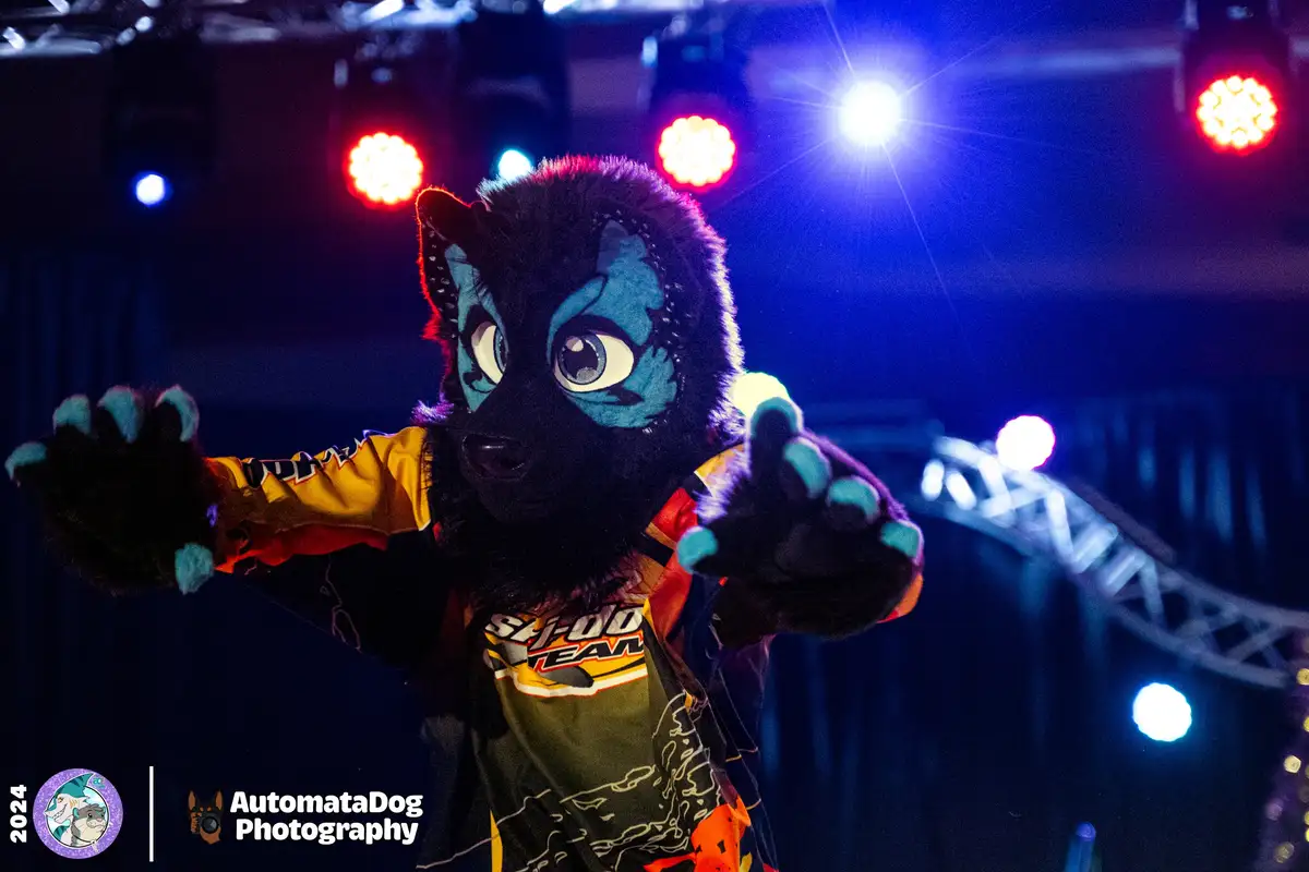 A person in a black and blue furry wolf costume with large eyes and clawed hands is on a stage under colorful stage lights…
