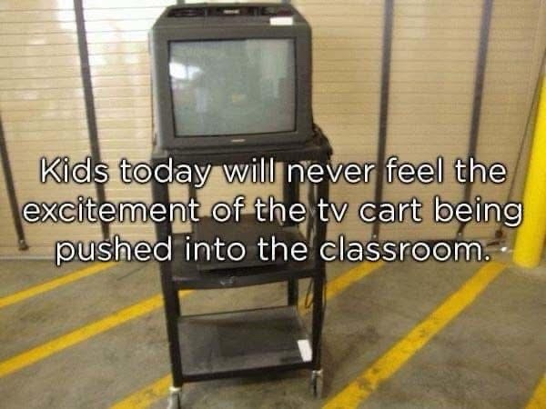 A black, wheeled TV cart with a bulky, old-style CRT television on top is parked in a warehouse or storage area with yellow…