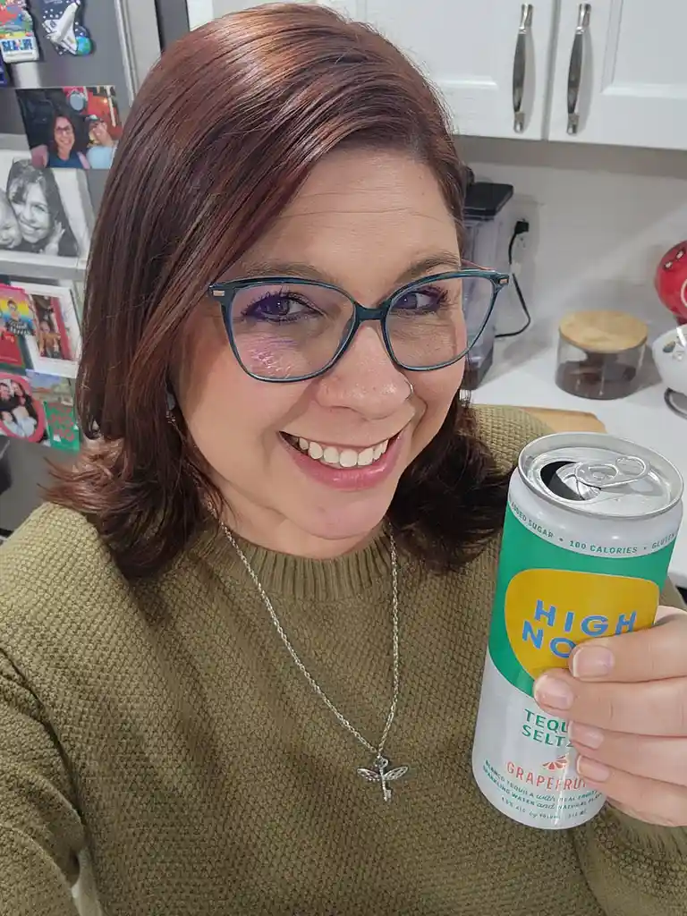 A woman with glasses and a nose ring smiles while holding an open can of 'High No' grapefruit tequila seltzer in a kitchen…