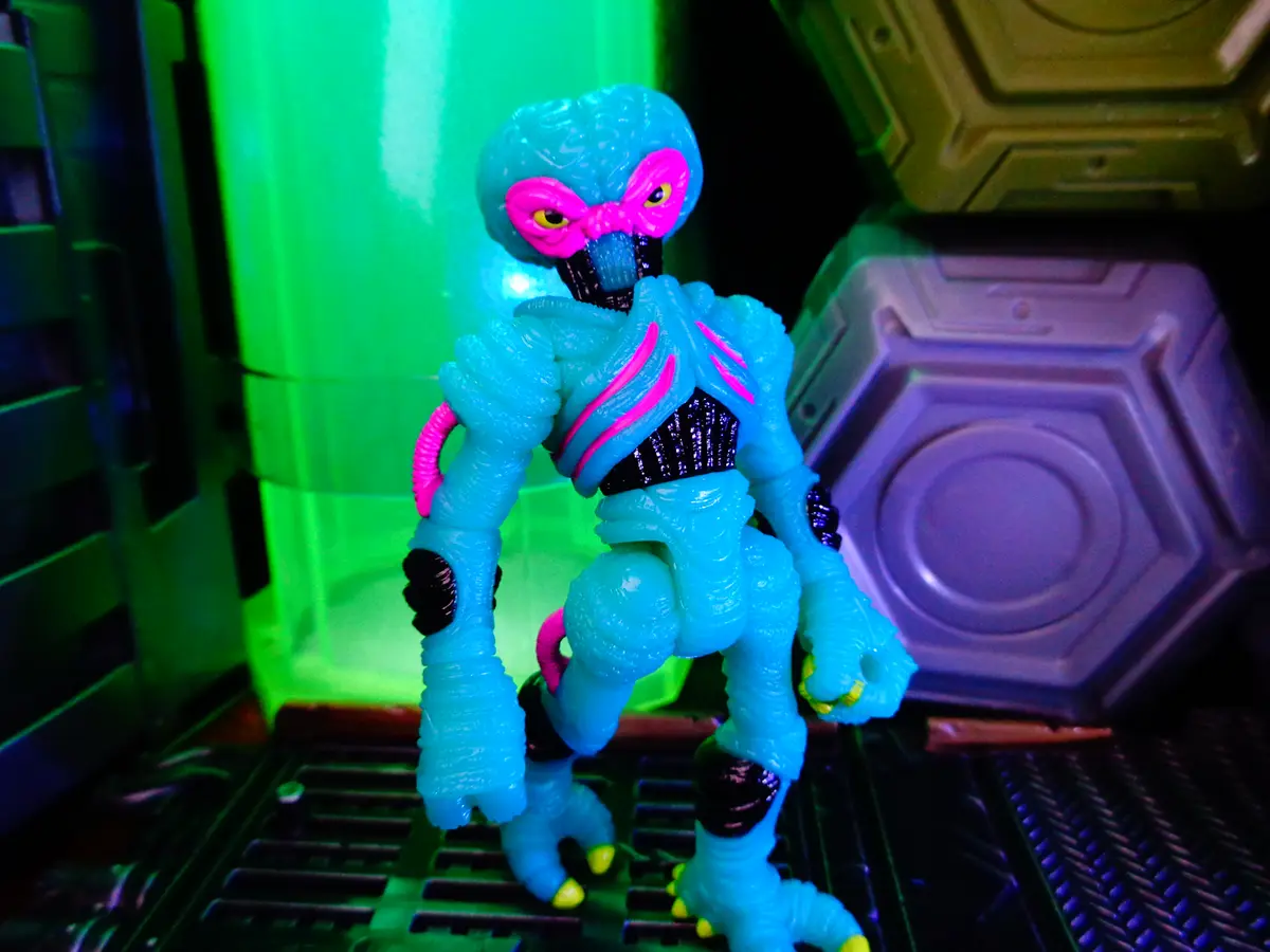 A blue alien action figure with pink facial markings and glowing yellow eyes stands in a sci-fi setting with green and…