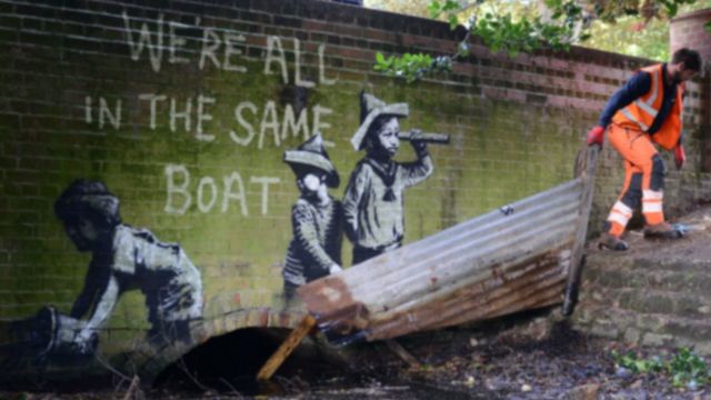 A mural depicting three children in a boat with the text 'WE'RE ALL IN THE SAME BOAT' is physically extended into the real…