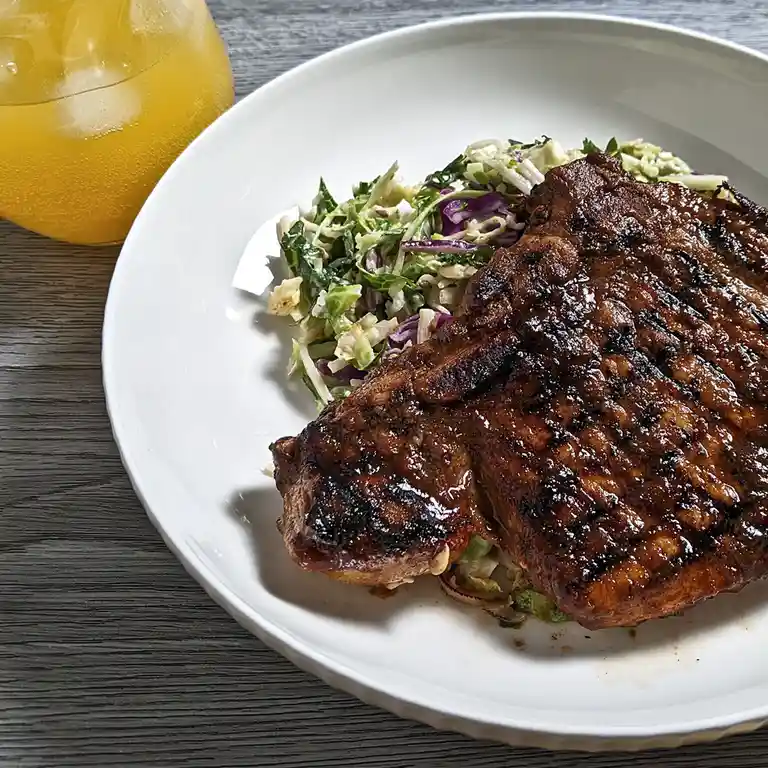 A close-up photo shows a white plate with a large, grilled, glazed piece of meat next to a colorful coleslaw salad, with a…