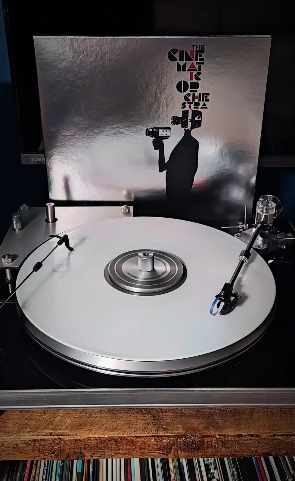 A vinyl record player with a white record spinning, its album cover behind it features a silver foil design with a black…