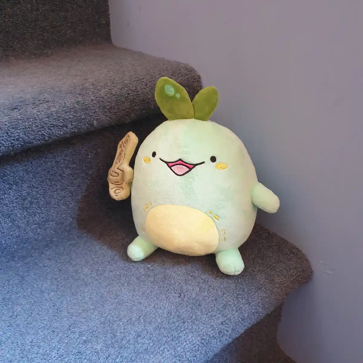 A light green, round plush toy with leaf-like ears and a cheerful expression sits on a carpeted staircase. It holds up a…