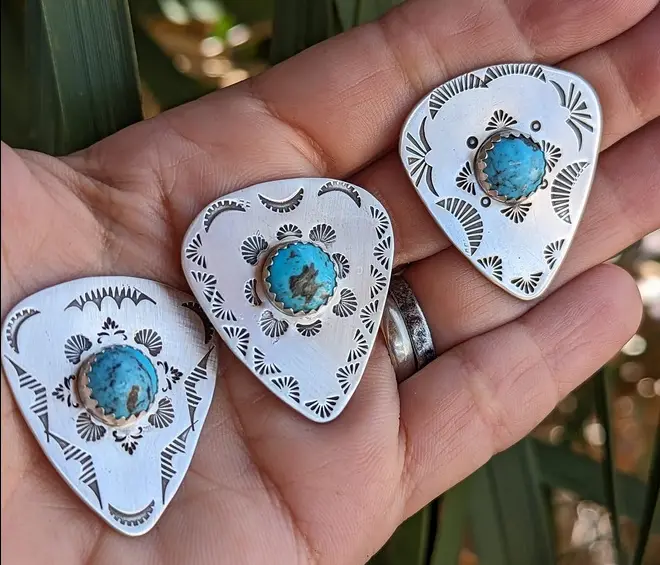 A hand holds three intricately designed silver rings shaped like guitar picks, each featuring a turquoise stone centerpiece…