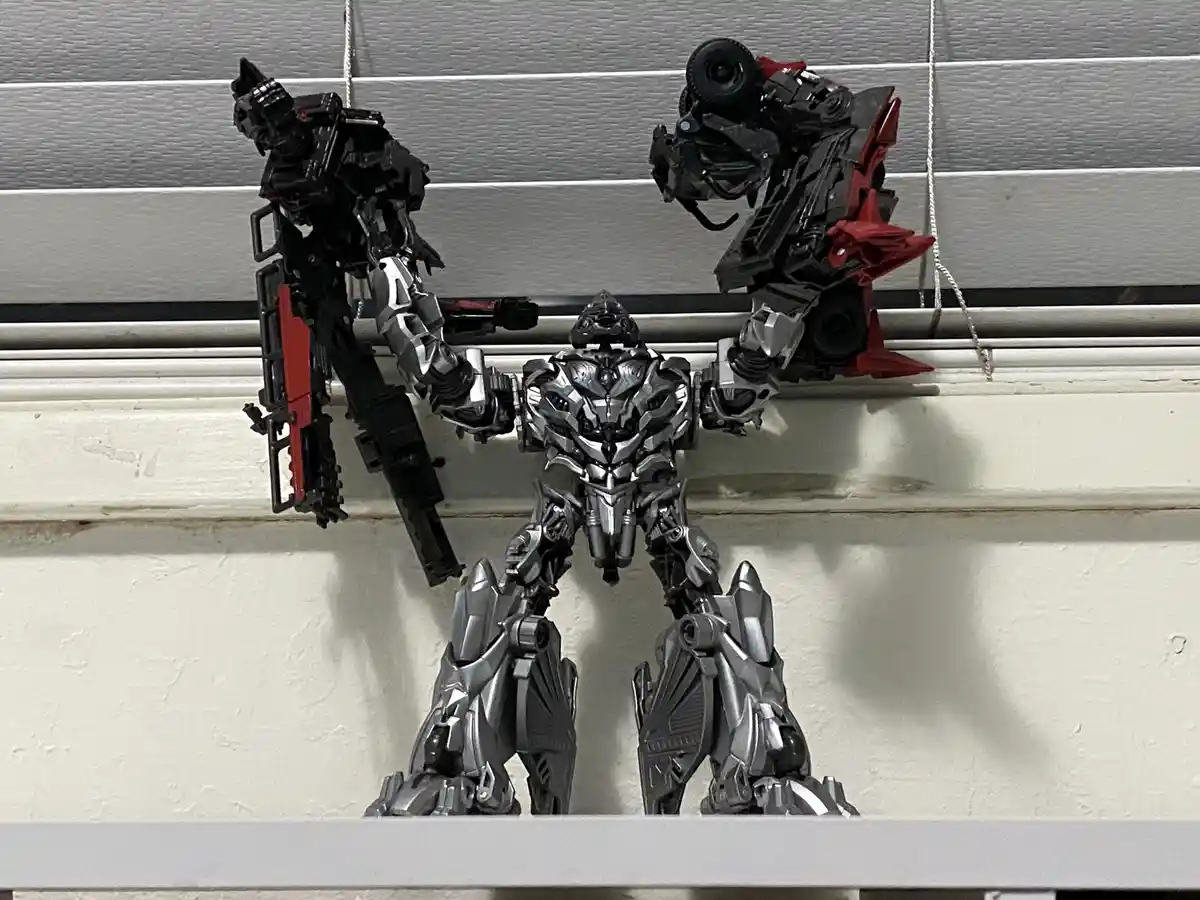 Three silver and black transformer action figures are posed on a shelf against a white wall with horizontal blinds. The…