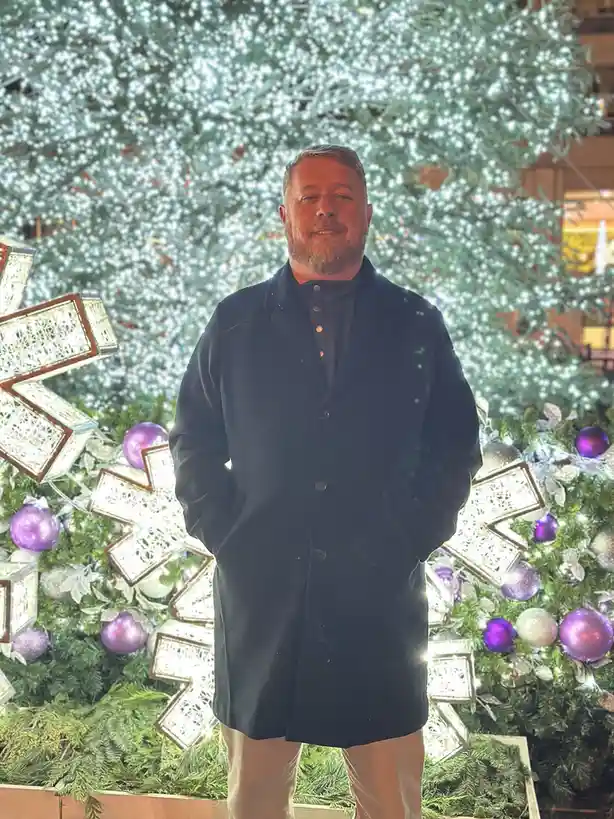 A man with a beard and dark coat stands in front of a brightly lit Christmas tree decorated with purple ornaments and large…