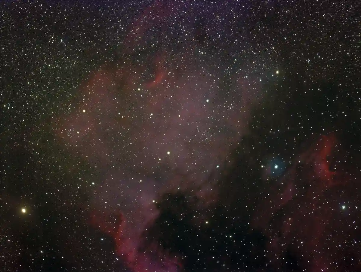 A deep-sky astrophotograph showing a starfield with numerous stars, glowing red and purple nebulae, and dark dust lanes…