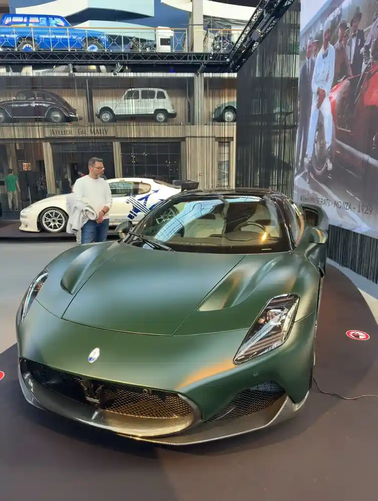 A matte green Maserati sports car is displayed at an auto show, with a man in a white shirt standing nearby. The background…