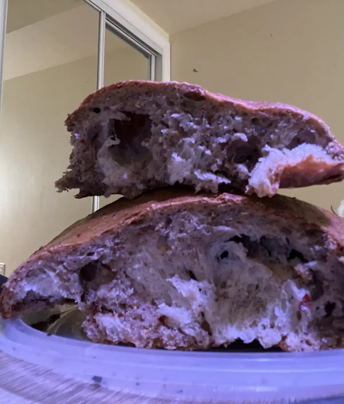 A close-up photo shows two halves of a rustic, seeded bread loaf stacked on a clear plastic container, revealing its airy…
