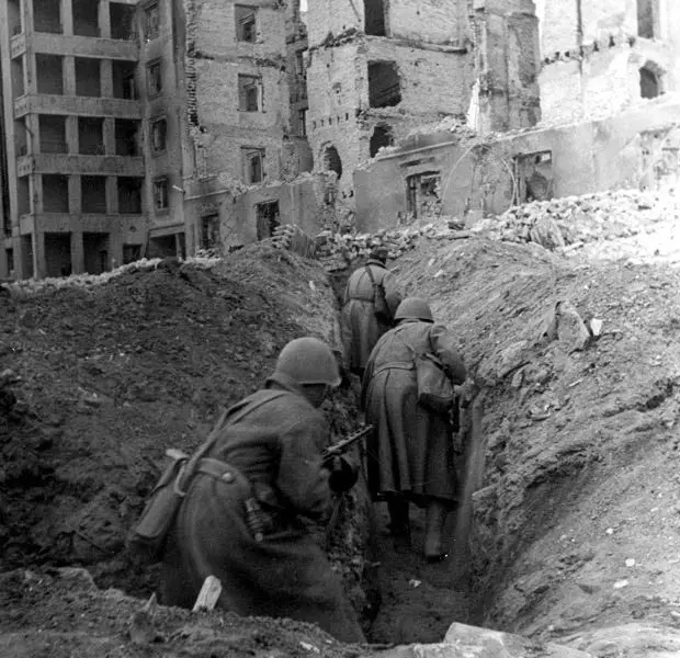 A black-and-white historical photo shows three soldiers in helmets and coats crawling through a muddy, narrow trench. Behind…
