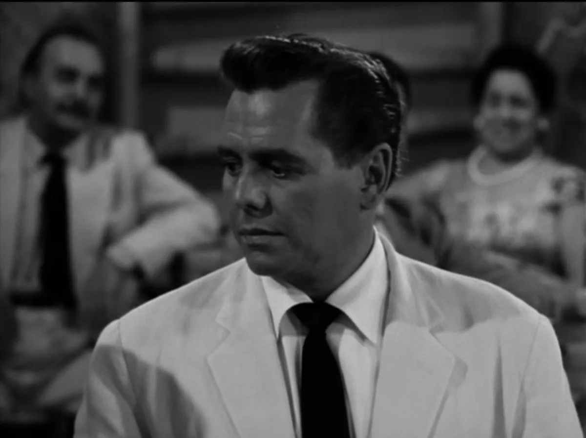 A black-and-white still of a man in a white lab coat and black tie looking concerned, with two blurred figures in the…