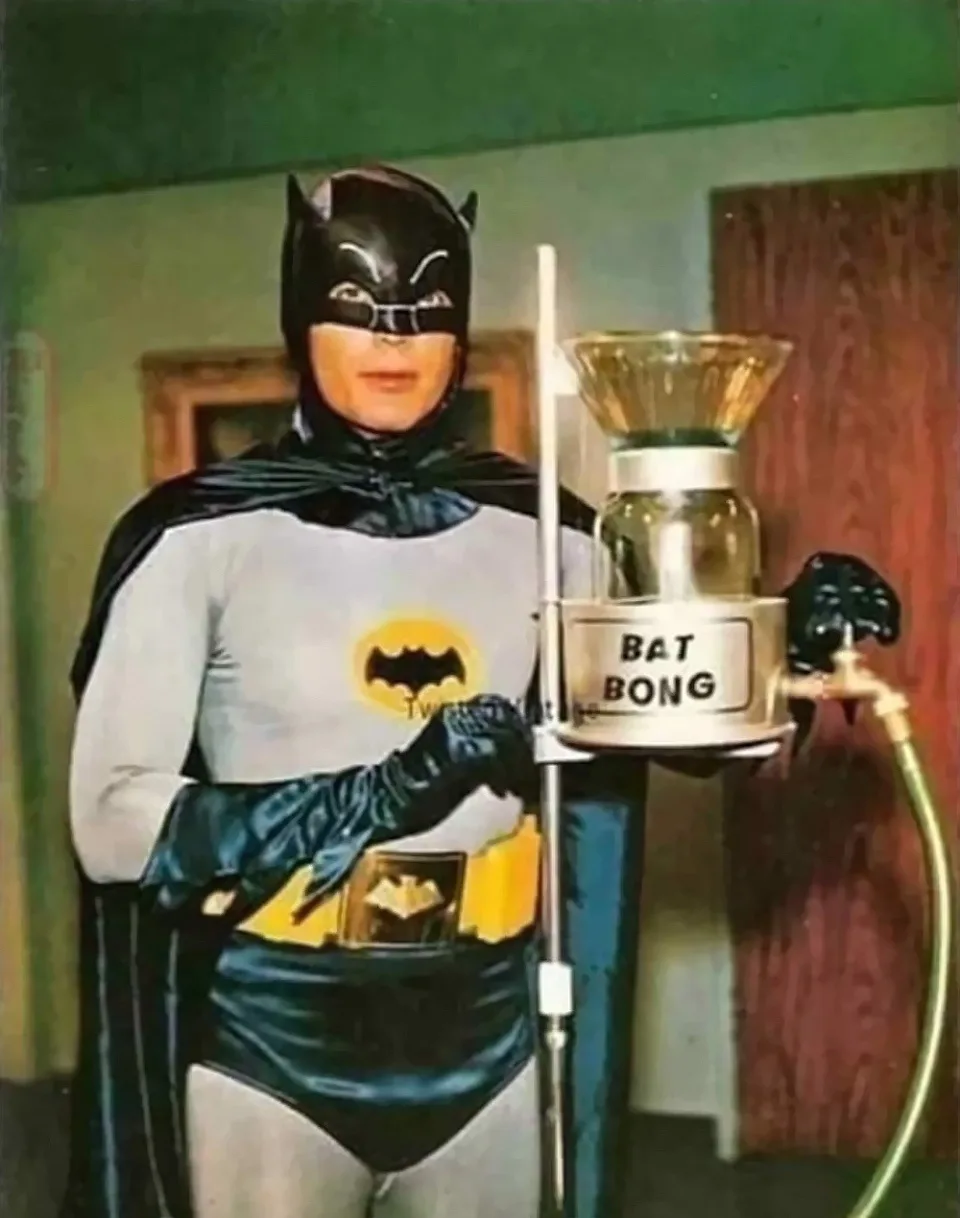 A person in a vintage Batman costume from the 1960s TV show stands holding a large glass bong mounted on a stand. The bong…