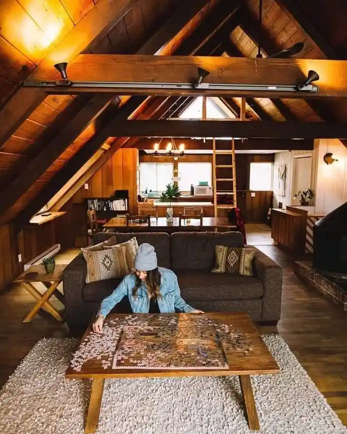 A woman in a grey beanie and denim jacket sits on a brown couch in a cozy, wood-paneled attic room, focused on assembling a…