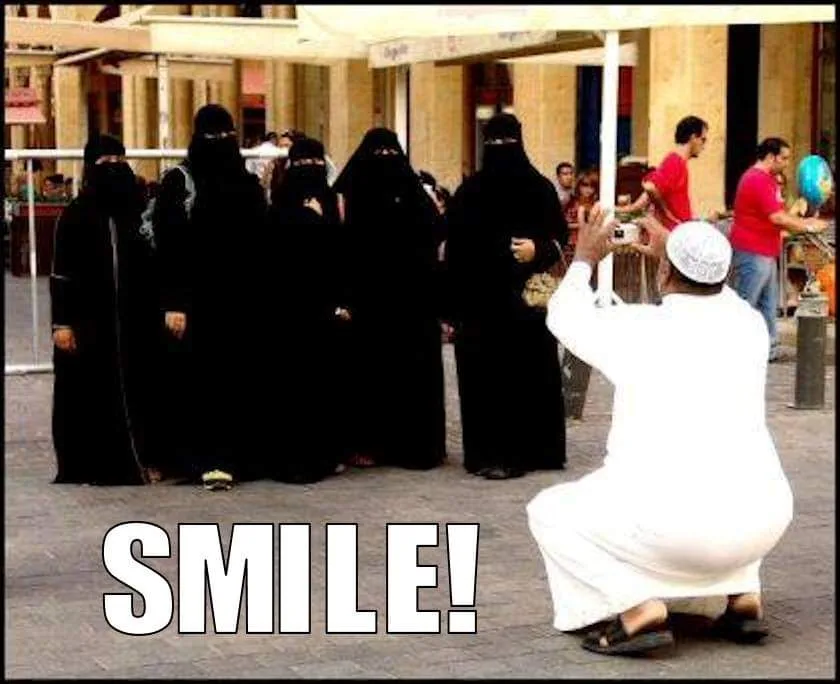 A man in white traditional clothing kneels and holds a camera toward a group of women dressed in full black niqabs, who…