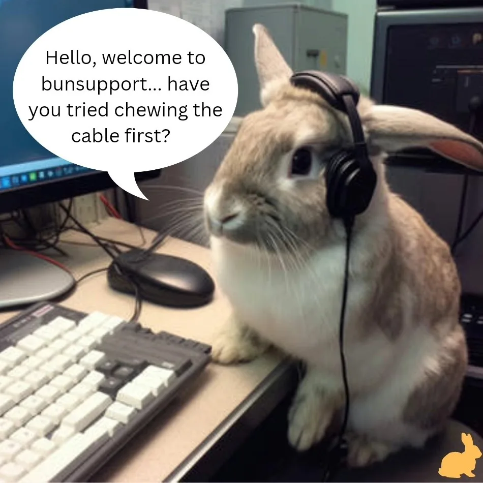 A light brown and white rabbit wearing black over-ear headphones sits upright on a desk next to a keyboard and mouse, with a…